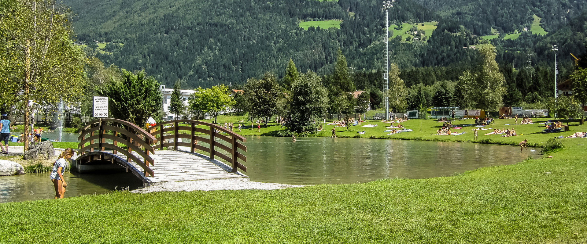 Romantischer Badesee mit einer kleinen Brücke, mitten im Grünen und von Bergen umgeben.