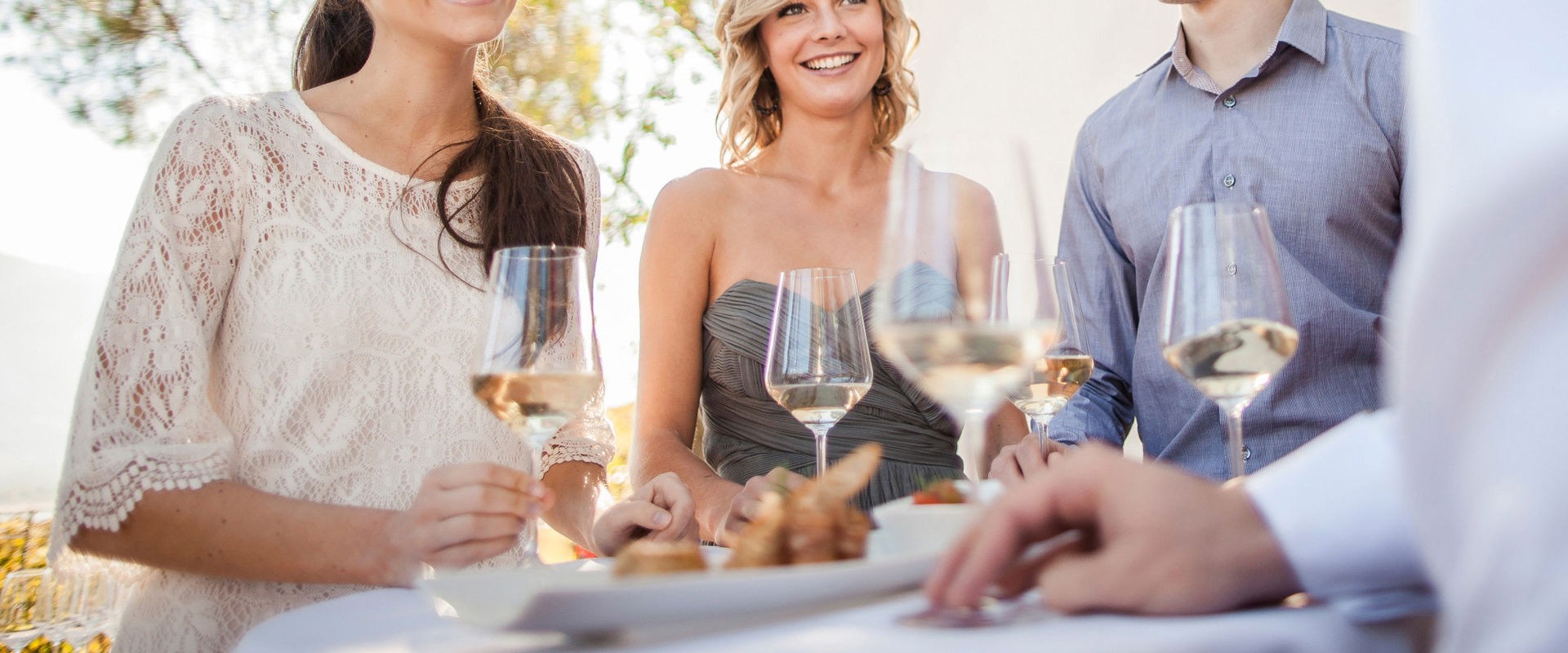 Gewürztraminer Junge Leute stehen an einem Tisch, mit einem Glas Weißwein in der Hand.