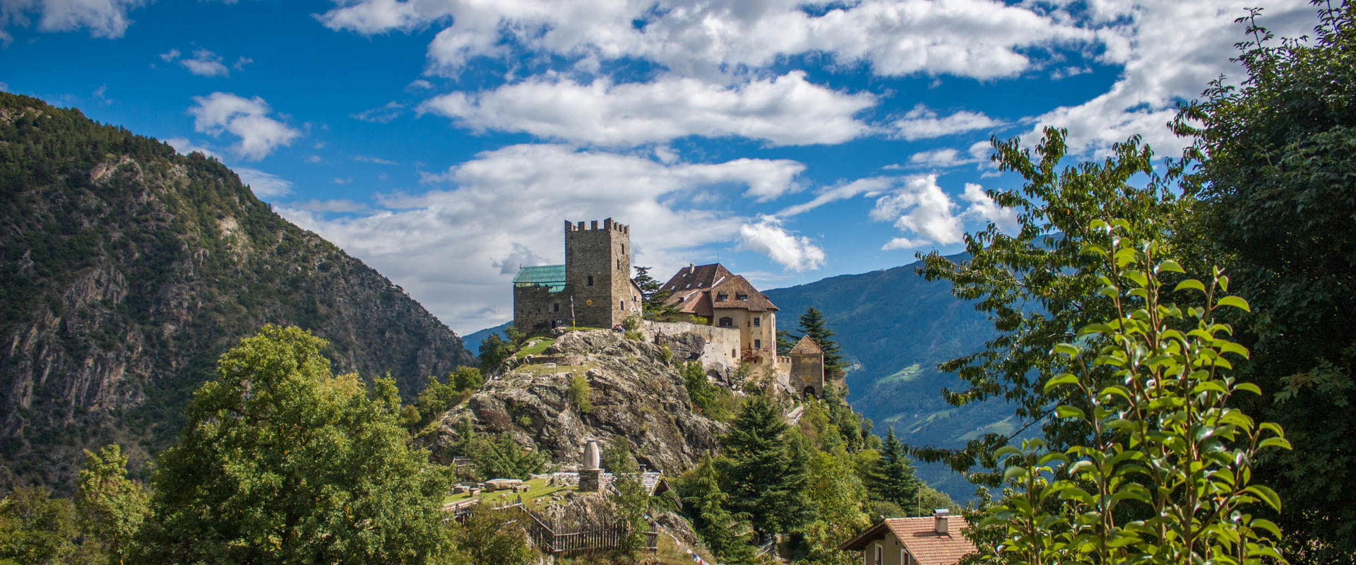 Schloss Juval Das Schloss Juval thront auf einem felsigen Hügel umgeben von Bäumen, Bergen, einem Haus und einer kleinen Hütte.