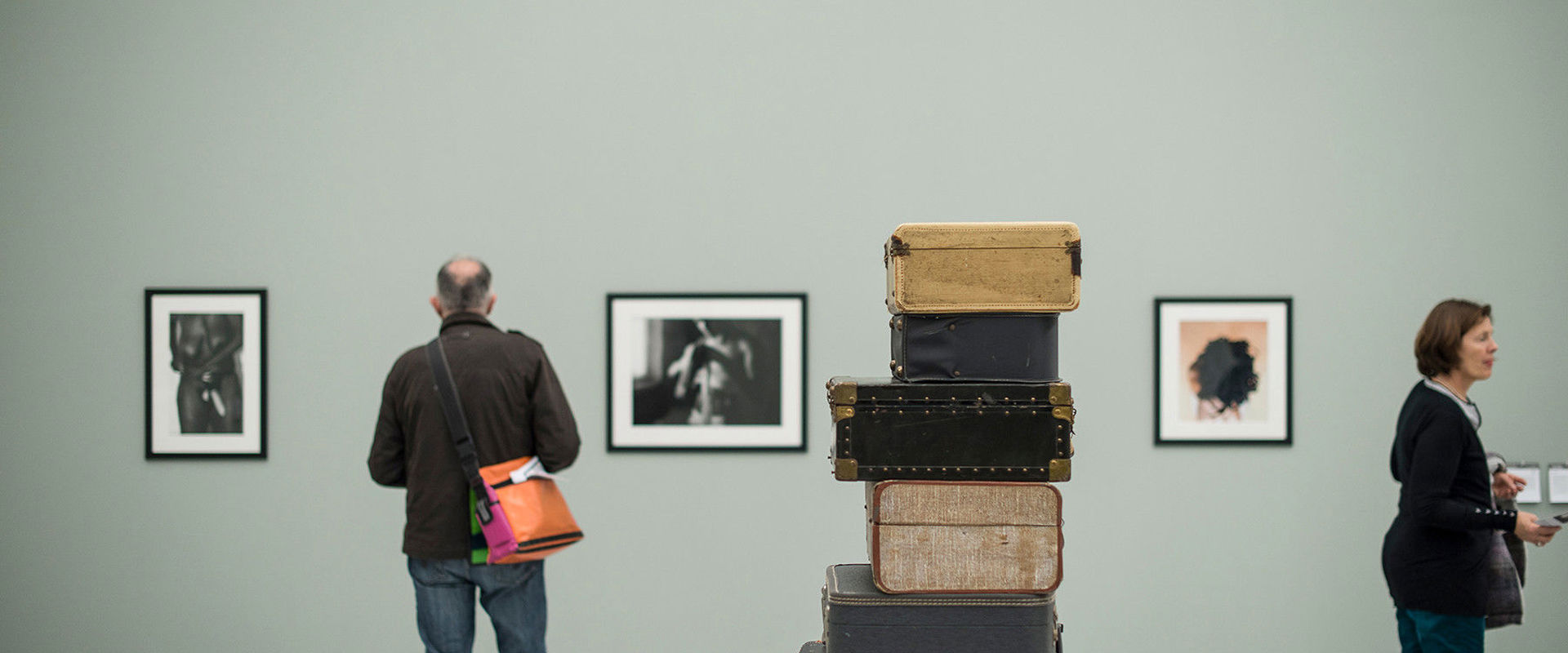 Mehrere alte Koffer liegen aufeinander mitten im Raum. An den Wänden hängen Bilder, die von 2 Besuchern begutachtet werden.