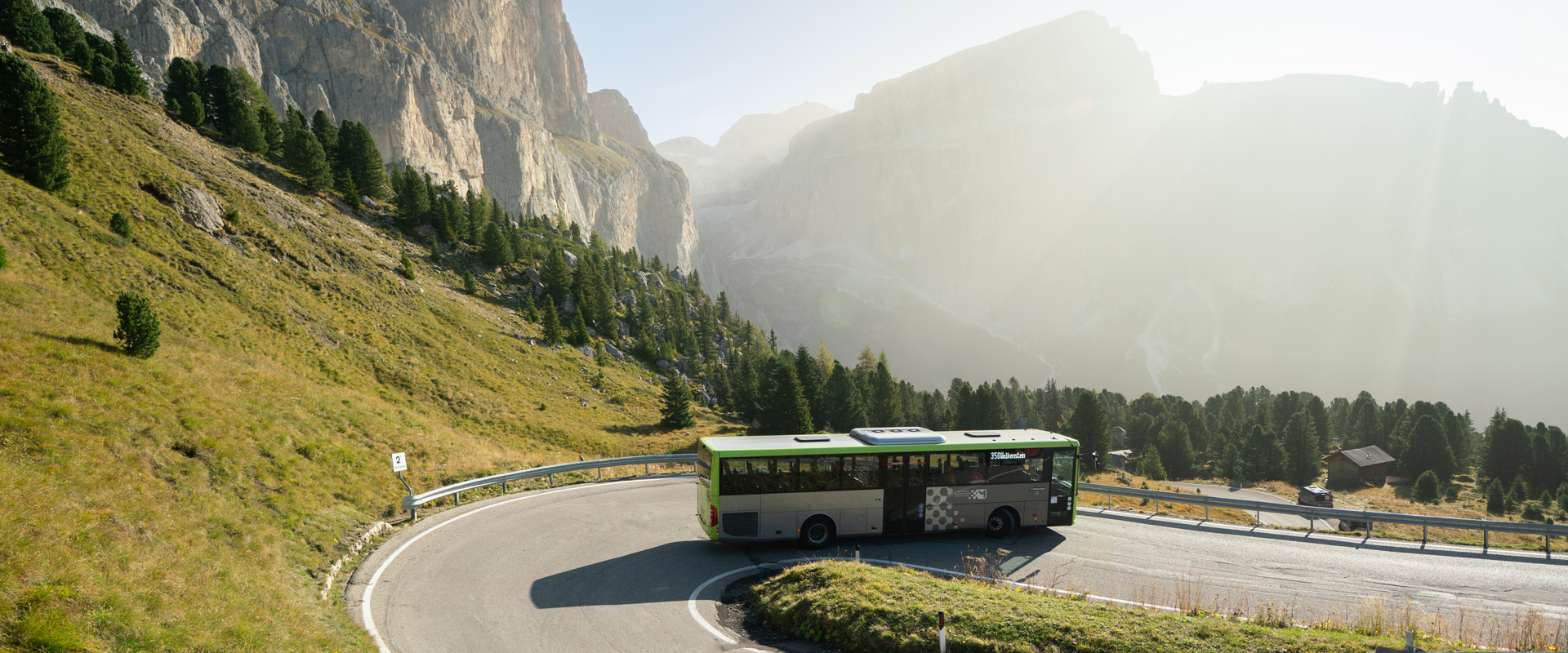 Mit dem Bus in die Dolomiten Bus fährt in einer Kehre bergab, dahinter sonnige Berglandschaft mit Wiese, Bäumen und Felswänden