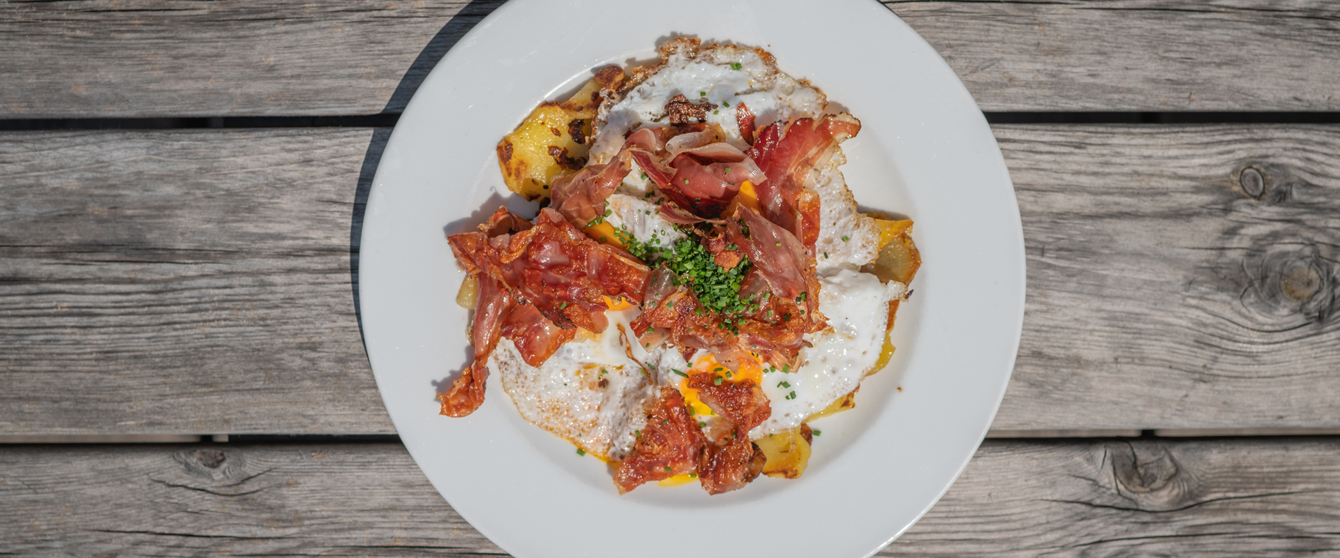 Teller auf Holztisch mit Röstkartoffeln, Speck und Eiern