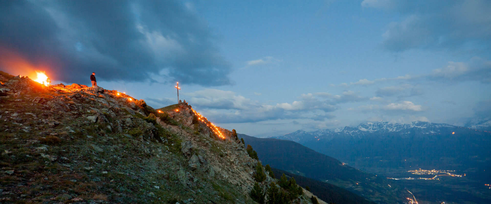 Herz-Jesu-Feuer am Berggipfel. Mann steht neben dem Gipfelkreuz und beobachtet das lodernde Herz-Jesu-Feuer.