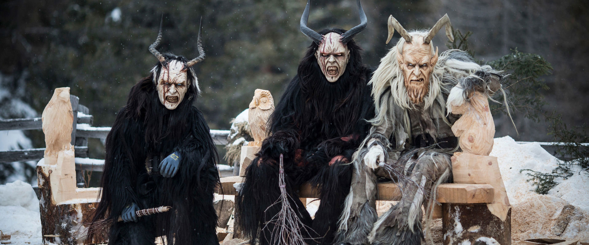 3 als Krampus verkleidete Personen sitzen auf einer Holzbank und schauen direkt in die Kamera.