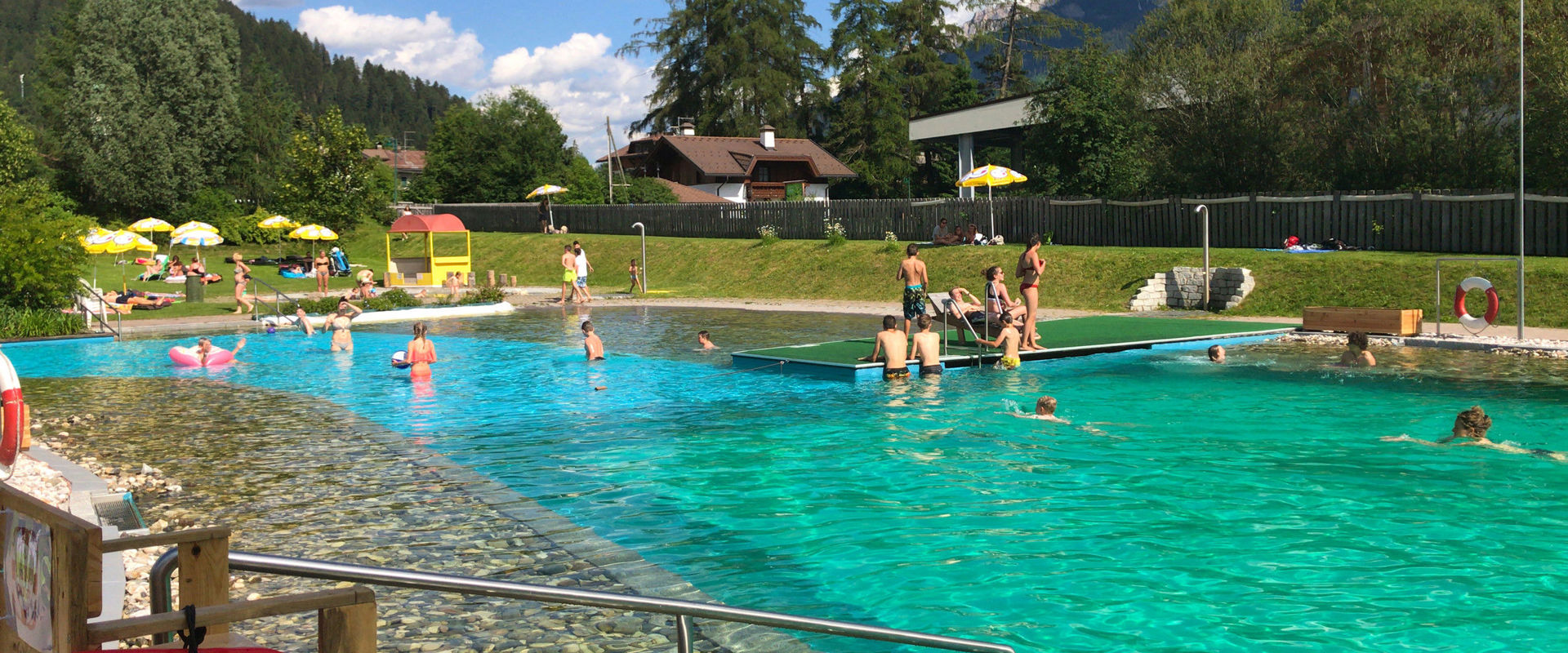 Badesee mit Kinderbecken, Steg, Duschen und Liegewiese. Im Hintergrund sieht man Häuser und Bäume.