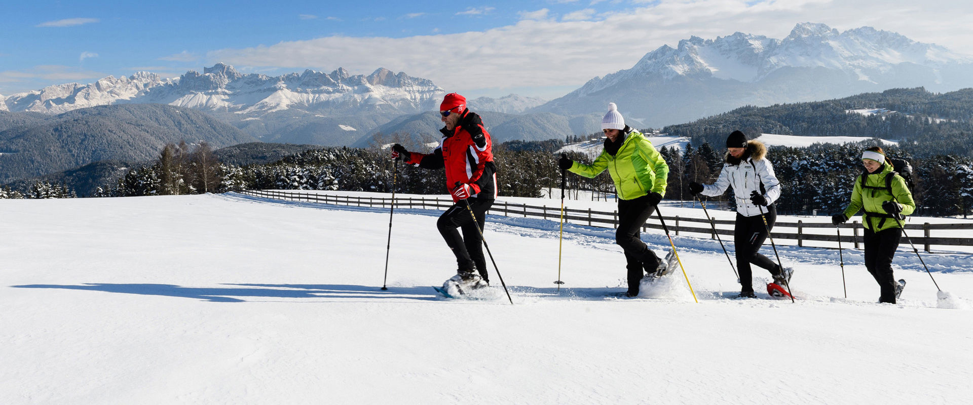 4 Schneeschuhwanderer im Tiefschnee auf einer eingezäunten Wiese. Im Hintergrund sieht man eine atemberaubende Berglandschaft.