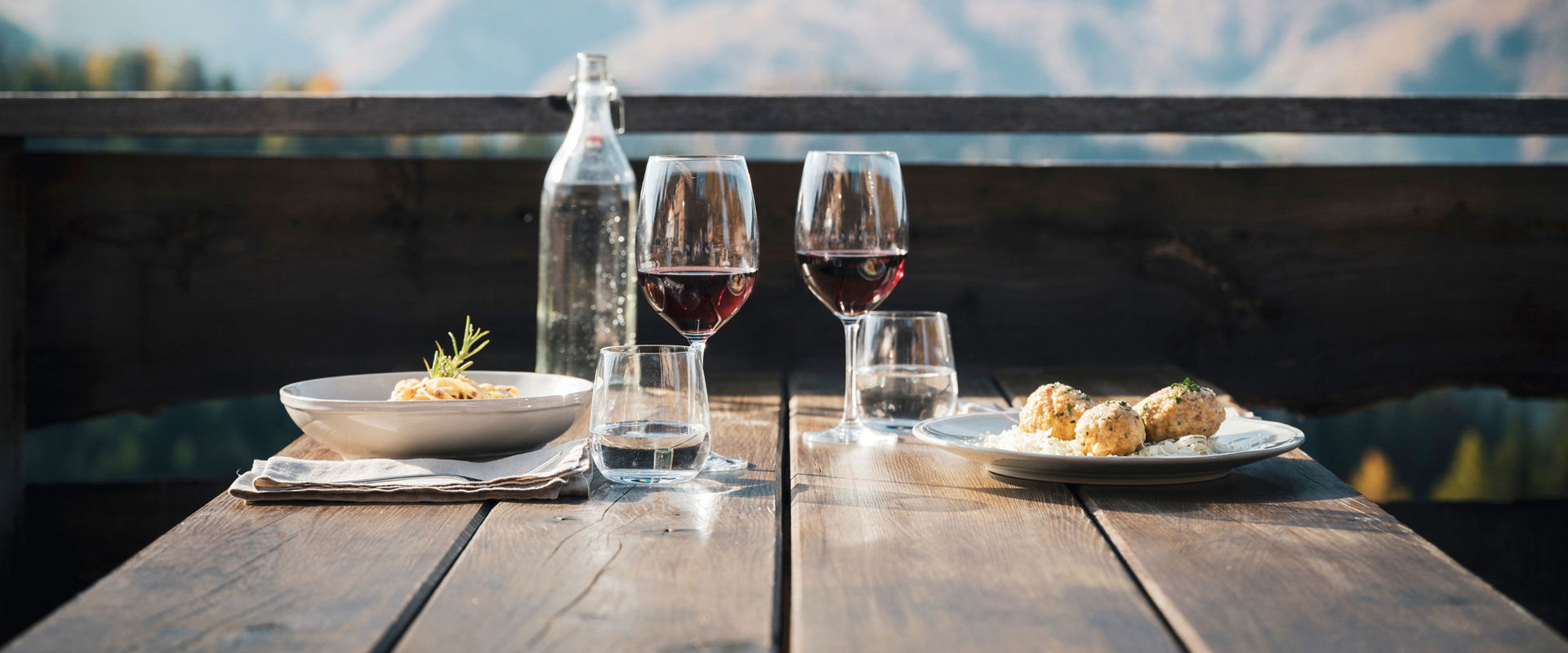 Essen im Sommer mit Dolomitenblick. 2 Teller und Weingläser auf einen Holztisch.
