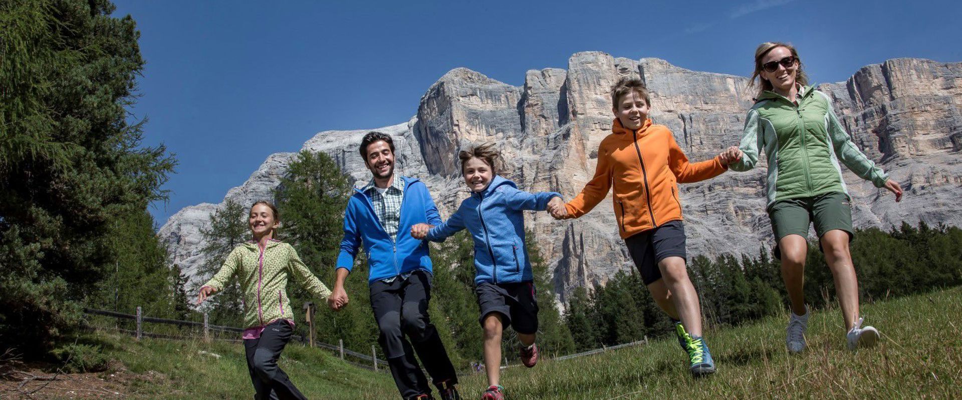 Familie hält sich die Hände und läuft auf einer Wiese. Im Hintergrund sieht man steile Felswände.