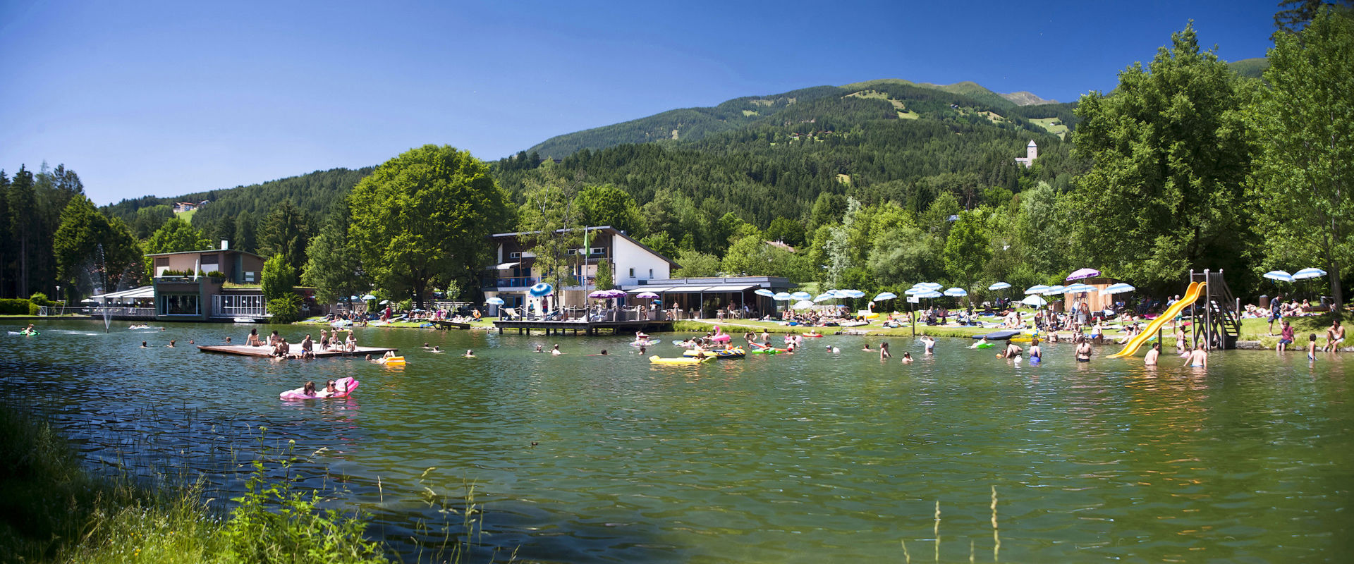 Badesee mit Restaurant, Steg, Floß, Rutsche und blau weißen Sonnenschirmen.