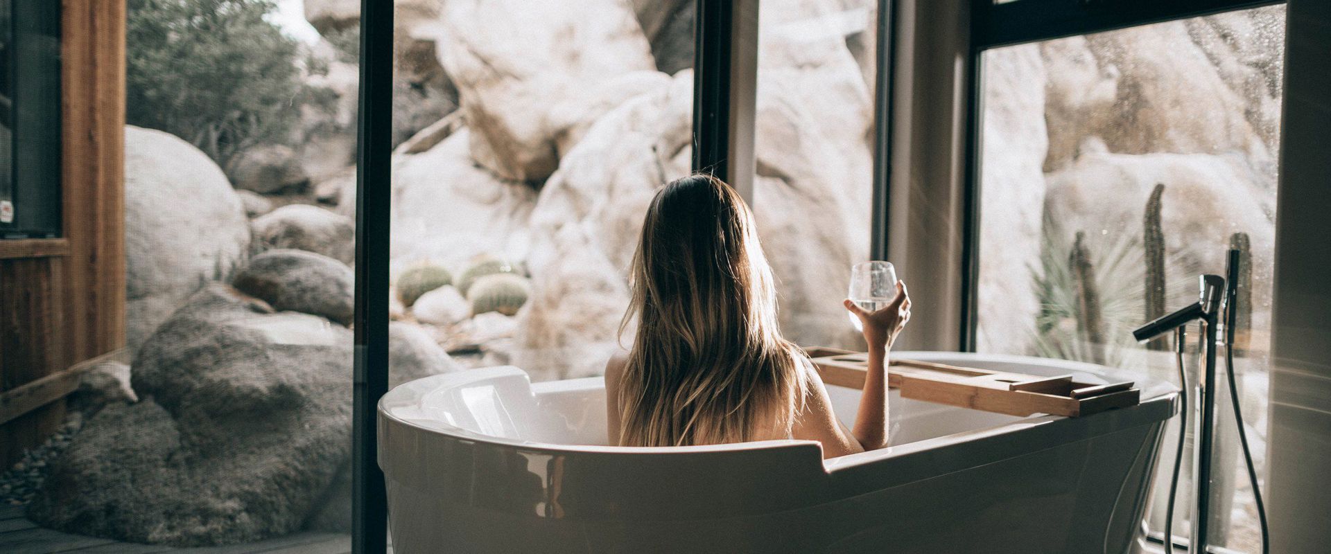 Frau sitzt in der Badewanne vor der verglasten Terrassentür und trinkt ein Glas Weißwein.