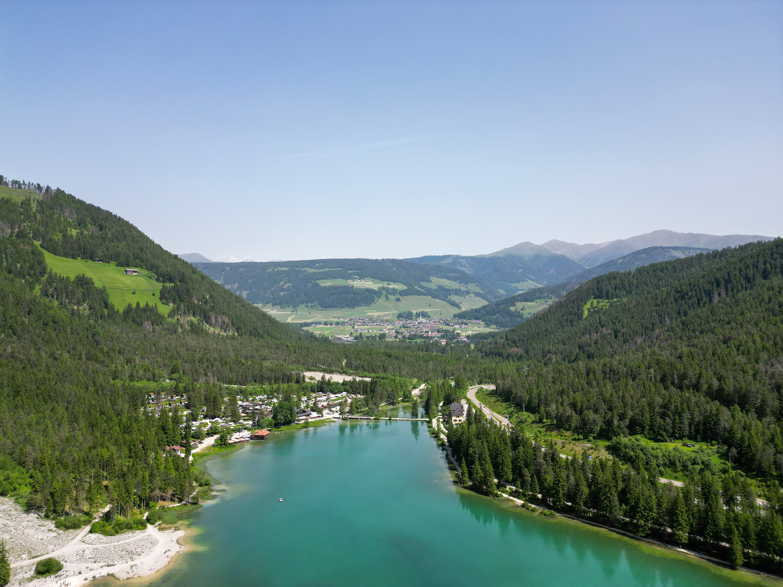 Von Wald und Campingplatz eingerahmter See, Berglandschaft mit Dorf im Hintergrund