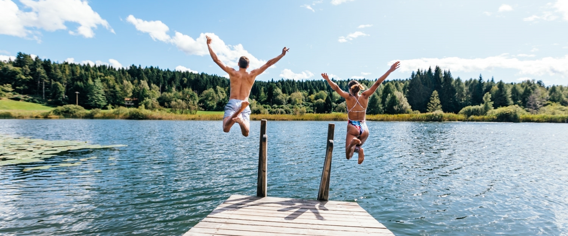 Eine Frau und ein Mann springen vom Holzsteg in den Fennberger See.