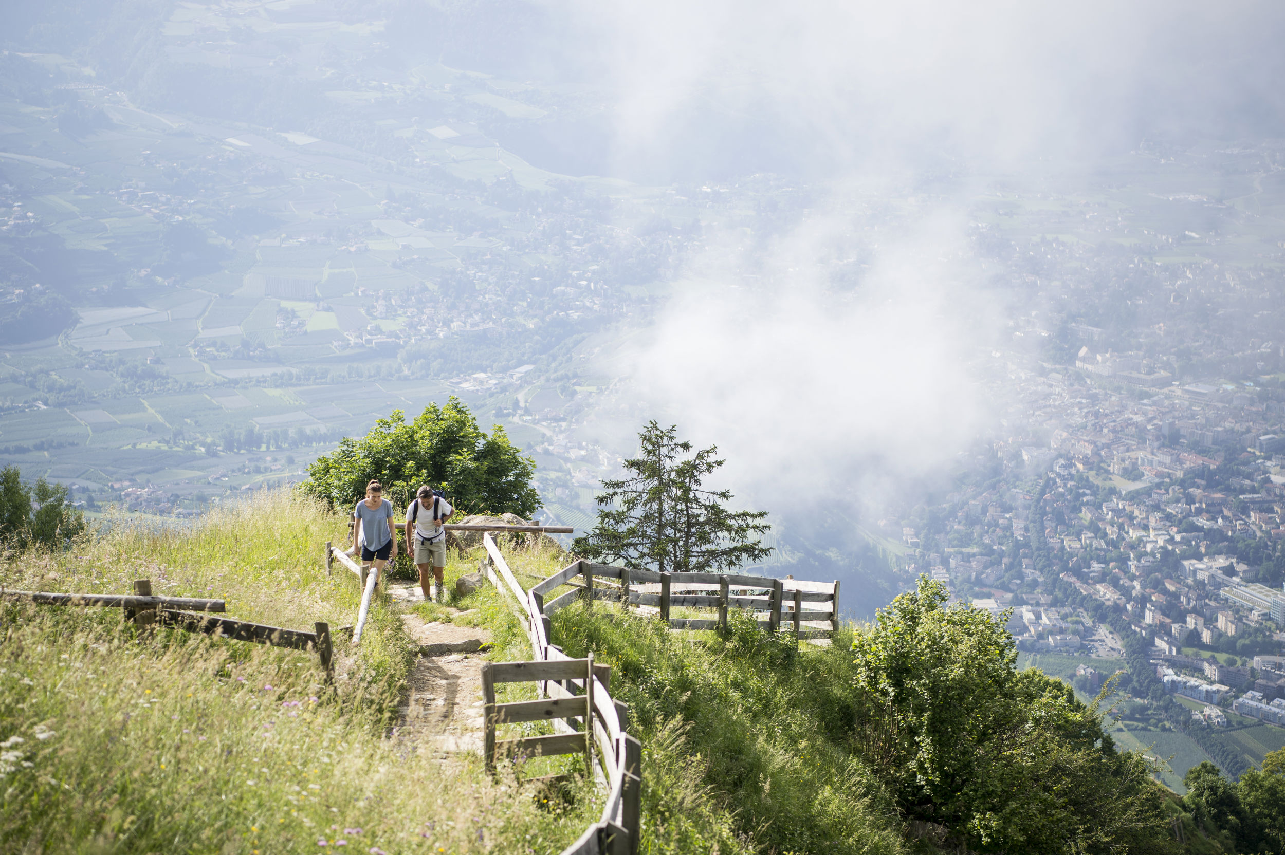 Zwei Wanderer gehen den Bergpfad hoch. Unten im Tal sieht man eine kleine Stadt.